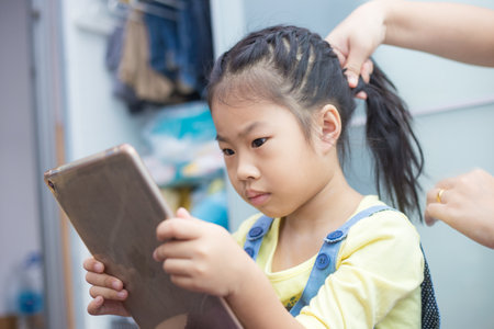 Adorable asian school girl use tablet computer while hair dressing by mom in houseの写真素材