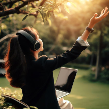 The back of an office girl who is using headphones to listen to music from a notebook with a joyful gesture in the garden.の素材