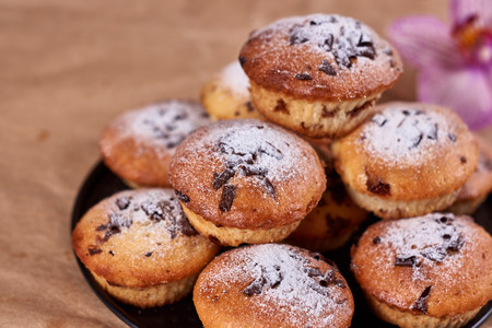 Cupcakes with chocolate chips with powdered sugar lie in a black baking sheet, on a kraft paper next to the orchid flower on a wooden background.の写真素材
