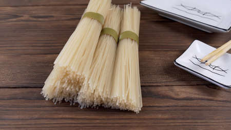 Dry unprocessed rice noodles and white square plate next to chopsticks on wooden backgroundの写真素材