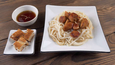 Rice noodles with fried chicken pieces in a white square plate next to fried bread and sauce on wooden backgroundの写真素材