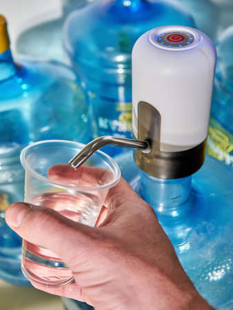 Portable electric pump for water bottle. A man pours water into a transparent plastic cup. Large bottle of pure water 19 liters with an automatic white pump in the background of other large bottlesの写真素材