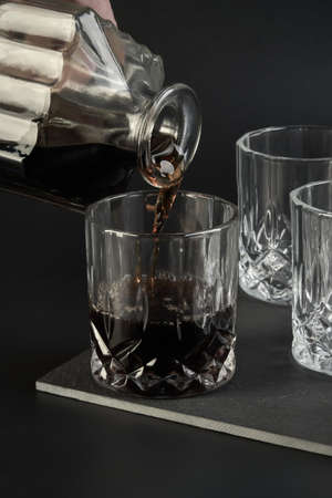 Someone is pouring cognac from a square decanter into a glass next to other empty cognac and whiskey...