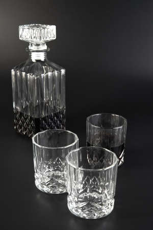 A square decanter of cognac stands next to cognac and whiskey glasses against a dark background....