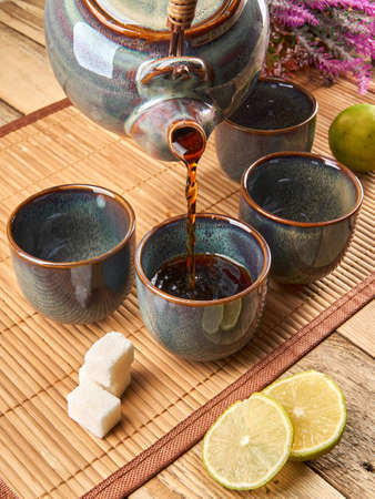 Chinese tea party. A ceramic teapot and fancy colored teacups sit on a mat next to sugar, limes and flowers. Someone pours tea from a teapot into a cupの写真素材