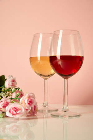 Two wine glasses are on a glossy table next to roses. Glasses with white and red wine on a pink backgroundの写真素材