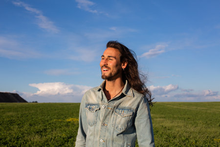 Young handsome man with long wavy hair in jeans shirt and jeans jacket standing in green wheat field...
