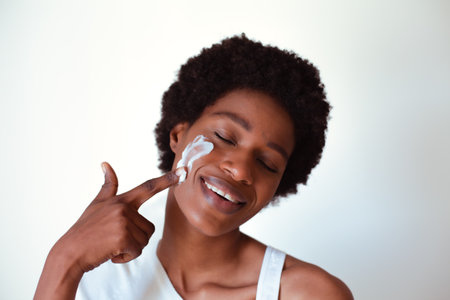 Portrait of a smiling young woman applying cream on her face against white backgroundの写真素材
