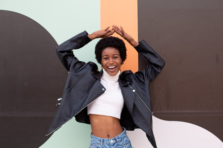 Beautiful african american woman with curly hair, wearing a black leather jacket and blue jeans, posing against colorful wall.の写真素材