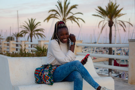 young happy and beautiful black afro American woman with dreadlocks in the city of Alicante at sunset listening to music with mobile phone smiling cheerful and relaxed sitting outdoorsの写真素材