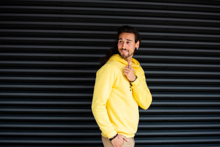 Portrait of a young man in a yellow hoodie standing against a black wallの写真素材