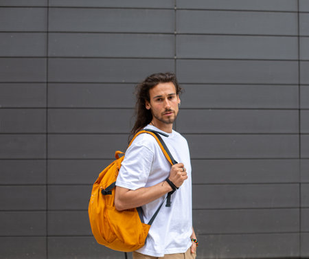 Portrait of a handsome young man with long curly hair, wearing a white T-shirt and yellow backpack, walking in the city.の写真素材