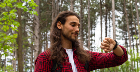 Handsome young man with long curly hair and beard in a red checkered shirt in the forest.