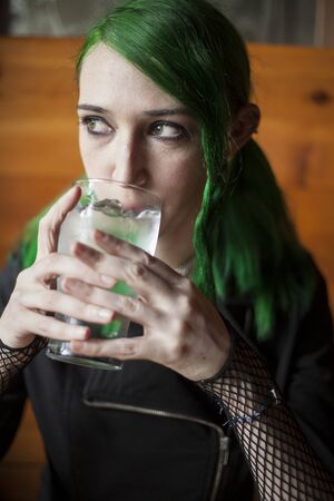 Portrait of a beautiful young woman with green hair and eyes drinking a glass of water at a pub.の写真素材