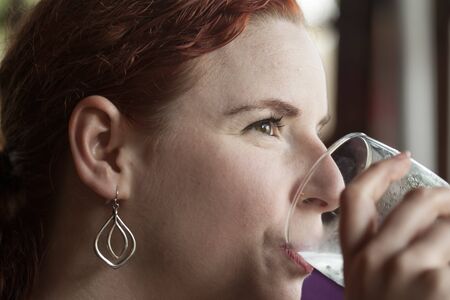 Beautiful young woman with red hair drinking water with ice from a pint glass.の写真素材