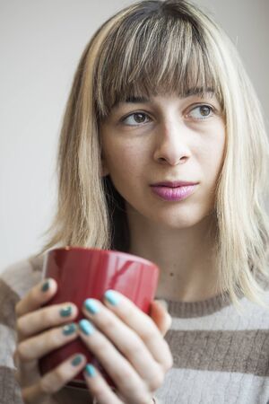 Portrait of a beautiful young woman with blonde hair drinking coffee from a red mug.の写真素材