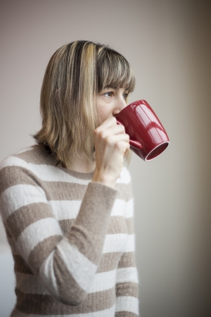 Portrait of a beautiful young woman with blonde hair drinking coffee from a red mug.の写真素材
