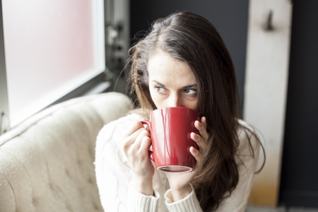 Beautiful young woman in a white sweater drinking coffee from a red cup.の写真素材