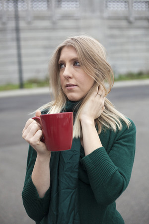 Photo of a very attractive blonde outside wearing a green top and drinking coffee from a red cup.の写真素材