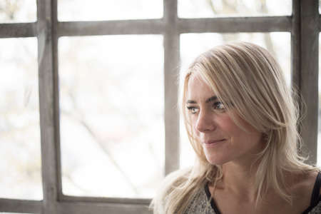 Photo of a very attractive blonde woman with beautiful blue eyes. She is standing in front of a window and wearing a gray sweater.の写真素材