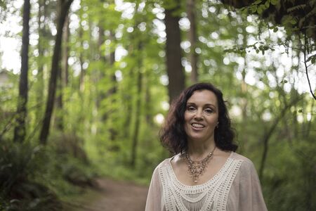 Photo of a very attractive 40-year-old woman with brown hair and eyes. She is standing in a wooded are and wearing a white top.の写真素材