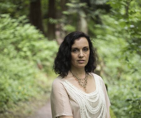 Photo of a very attractive 40-year-old woman with brown hair and eyes. She is standing in a wooded are and wearing a white top.の写真素材