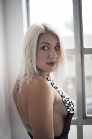 Photo of a very attractive blonde woman with beautiful blue eyes wearing a sleeveless top and standing in front of a window.の写真素材