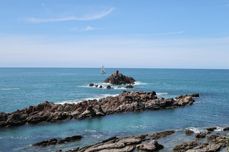 Rocky Islet Lighthouse Beside Passing White Sailboatの写真素材