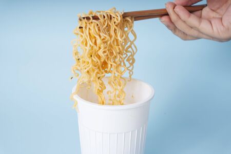 Hand of person using wooden chopstick eating instant noodle from a cup over blue background.の写真素材