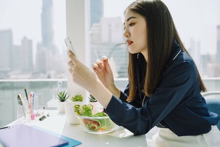 Beautiful asian young thai woman long dark hair enjoy eating salad bowl at office desk. Looking the phone.の写真素材
