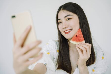 Beautiful asian young thai woman long dark hair holding piece of watermelon taking selfie with smartphone.の写真素材