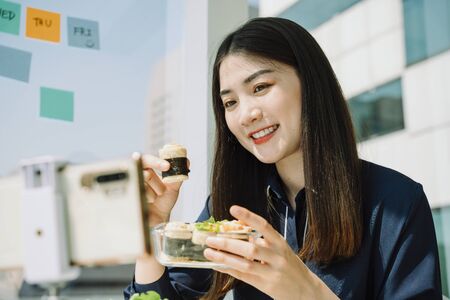 Beautiful asian thai long dark hair woman doing healthy food vlog with smartphone at office desk. City background.の写真素材