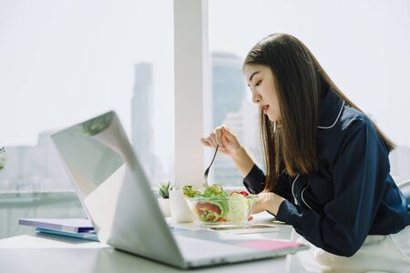 Beautiful asian young thai woman long dark hair enjoy eating salad bowl at office desk.の写真素材