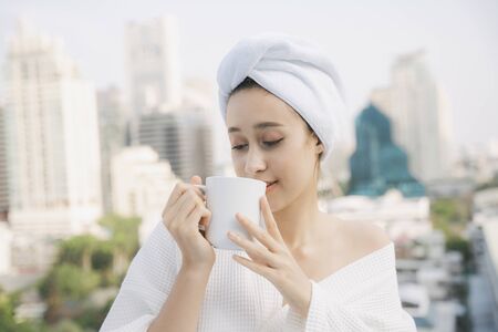 Young woman in white bathrobe enjoy hot coffee at apartment balcony over city background.の写真素材