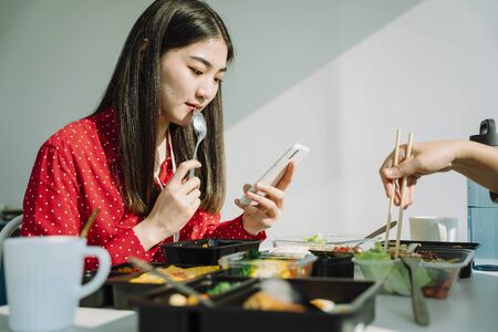 Beautiful young asian thai woman in red shirt eating and looking phone. Bad habit.の写真素材