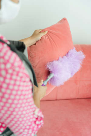 Maid cleaning sofa pillow couch with whisk broom shuttlecock in living room. Covid-19 Coronavirus prevention.の写真素材