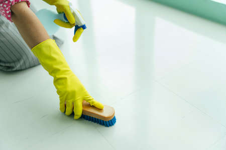 Maid wearing gloves cleaning floor with scrubbing brush and detergent spray bottle.