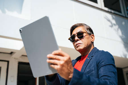 Old man in red t-shirt and sunglasses look at his reflection from the tablet and proud in his...