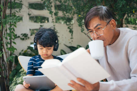 Little boy wearing the headphone to listen the music and read the e-book from tablet while the old man drink the coffee and read the book.の写真素材