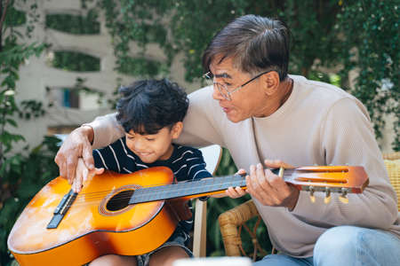 Old man with gray hair teach his grandson play the guitar in the backyard of his house.の写真素材