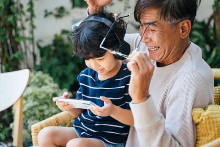 Little boy sitting with his grandpa on wicker chair in backyard and play the game on smartphone,...