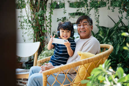 Little boy sitting with his grandpa on wicker chair in backyard and play the game on smartphone,...