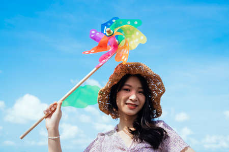 Happy cute girl wearing brown hat hold the colorful windmill in windy day under blue sky and white cloud in summer.の写真素材