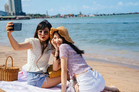 Two girls sitting on the beach at the sea in sunny day, take a selfie together and making duck face.の写真素材