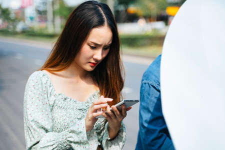 Angry asian woman start calling mechanic with phone to come here and fix her car. Young woman using mobile phone while looking at broken down car on street.の写真素材