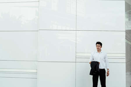 Mature asian business man on white formal shirt holding suit on arm standing at white wall, corner...