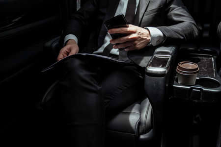 Businessman on black suit sittin in a car holding smartphone on the way to meeting.の写真素材