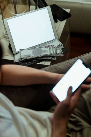 Close-up image of male hands using smartphone at home with white blank space on laptop in background. Man hand holding using mobile phone sitting on sofa.の写真素材