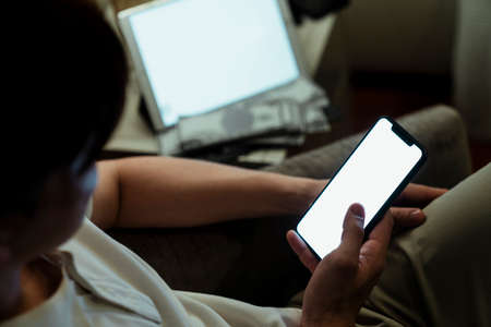 Close-up image of male hands using smartphone at home with white blank space on laptop in...