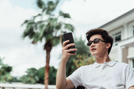 Young asian guy on white shirt wearing sunglasses using mobile phone at resort villa.の写真素材